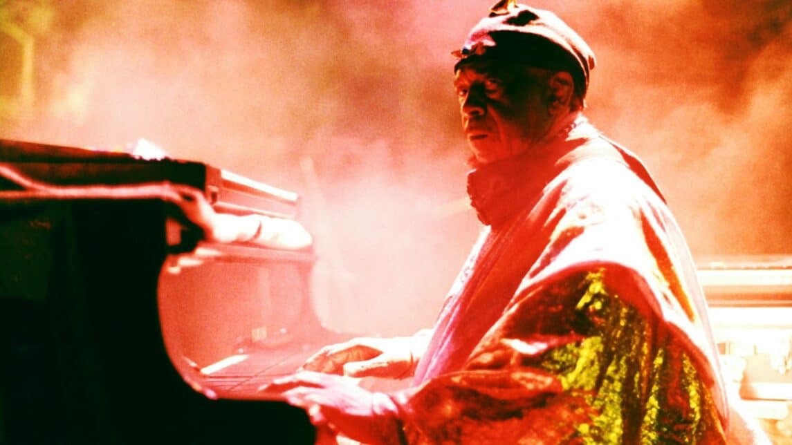 A film still from Sun Ra: Do the Impossible shows a Black man dressed in a red and metallic gold outfit seated at a piano. He looks to the side, into the camera. A warm colored fog surrounds him, filling the space with a hazy glow.