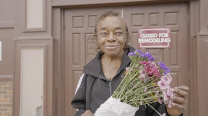 Film participant holds a bouquet of flowers. A sign with text: "Closed for Remodeling" hangs on a building in the background.
