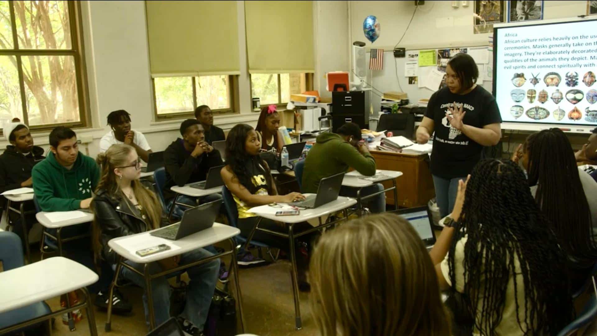 A film still from Teaching America shows a classroom of majority Black students students seated at desks arranged to form an aisle down the center of the room. The students’ attention is focused on a Black woman who stands in this aisle. She wears a black T-shirt that reads: “Central High NAACP” with a graphic of Africa in the center.