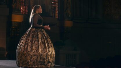 A film still from Tessitura shows a Black woman standing on the stage of a grand room, presumably singing. Her elbows are bent with hands in front of her. She wears a black sheer long-sleeved top and a black floor length skirt with a gold pattern.