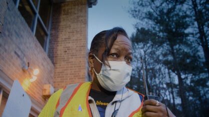 A film still from The Devil is Busy Shows an older Black woman wearing a face mask, her gaze looking at something off screen. She wears a neon yellow and orange vest over a grey zip-up hoodie. Held in front of her shoulder is a walkie-talkie.