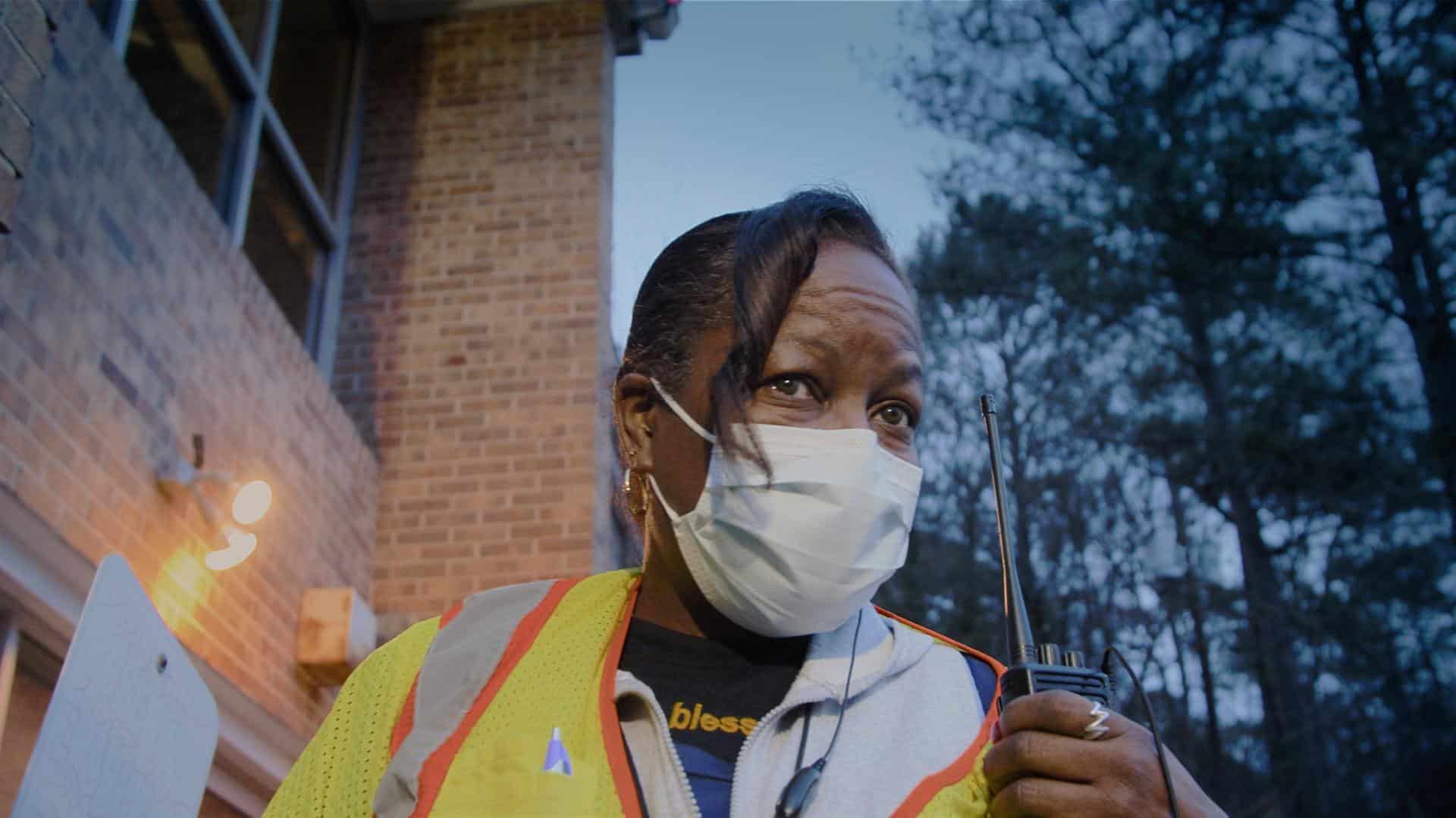 A film still from The Devil is Busy Shows an older Black woman wearing a face mask, her gaze looking at something off screen. She wears a neon yellow and orange vest over a grey zip-up hoodie. Held in front of her shoulder is a walkie-talkie.