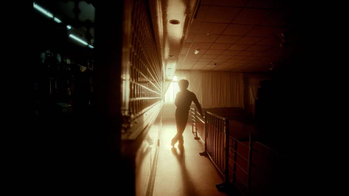 A film still from The Great North shows a silhouette standing in the center of a dark room, illuminated solely by a window whose curtain is pulled open. The silhouette is positioned between a railing and a wall that extends to the camera. The other side of the wall reveals a dimly lit room with indiscernible items.