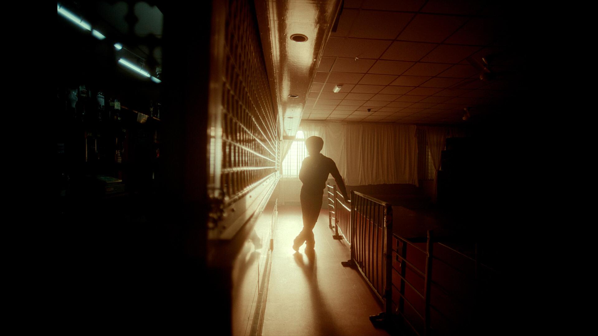 A film still from The Great North shows a silhouette standing in the center of a dark room, illuminated solely by a window whose curtain is pulled open. The silhouette is positioned between a railing and a wall that extends to the camera. The other side of the wall reveals a dimly lit room with indiscernible items.