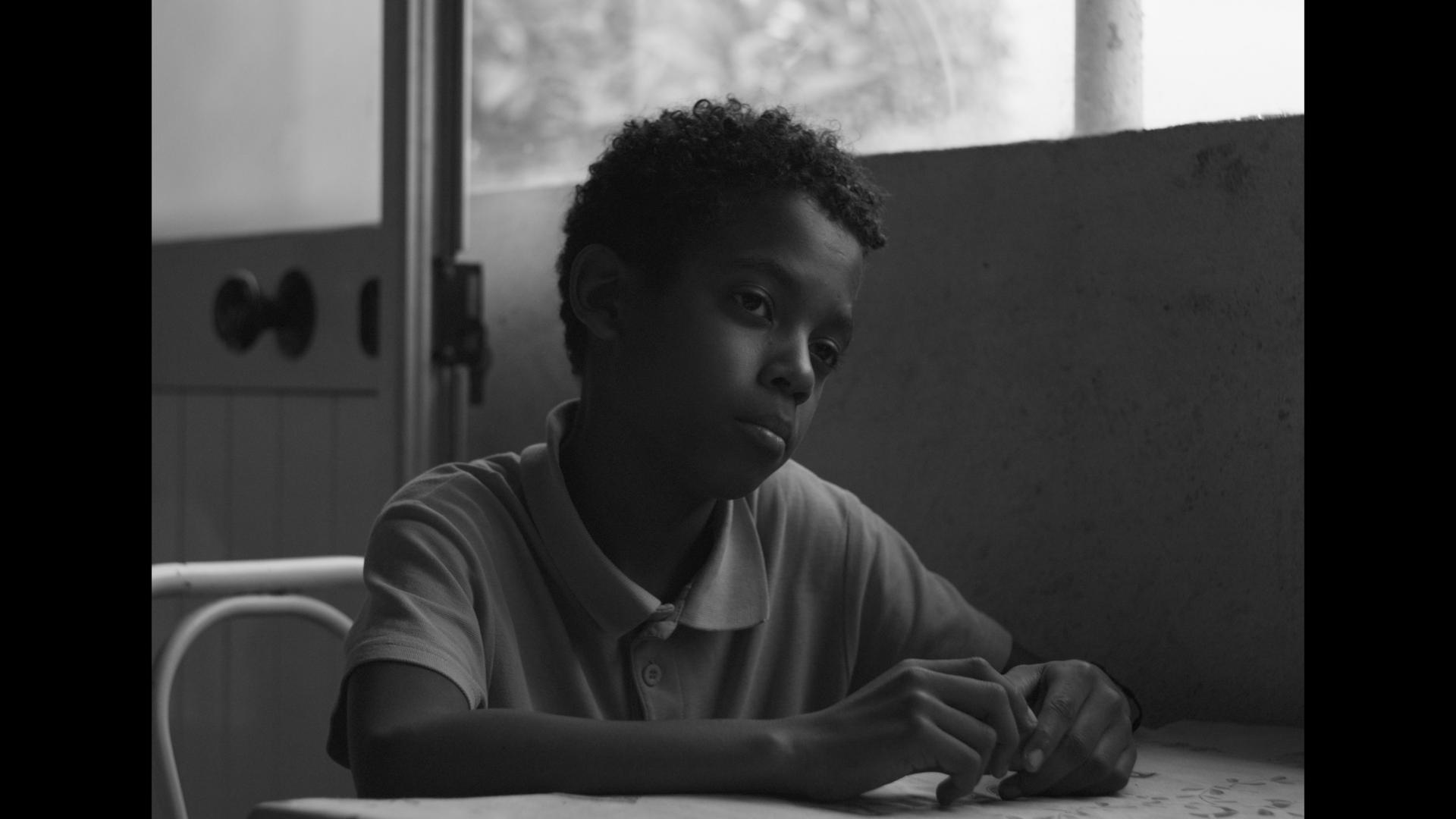 A black-and-white film still from The Last Harvest shows a disheartened young Black boy sitting at a table. His gaze is fixed off screen as his forearms rest on the table, hands touching.