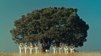 A film still from The River shows a wide shot of a large tree against a clear blue sky. Under the tree stands a woman next to a black and white board in which a child sits in front of. To each side of the child stand four people dressed in white head wraps, white robes with golden crosses in the center, and an orange and gold stole.