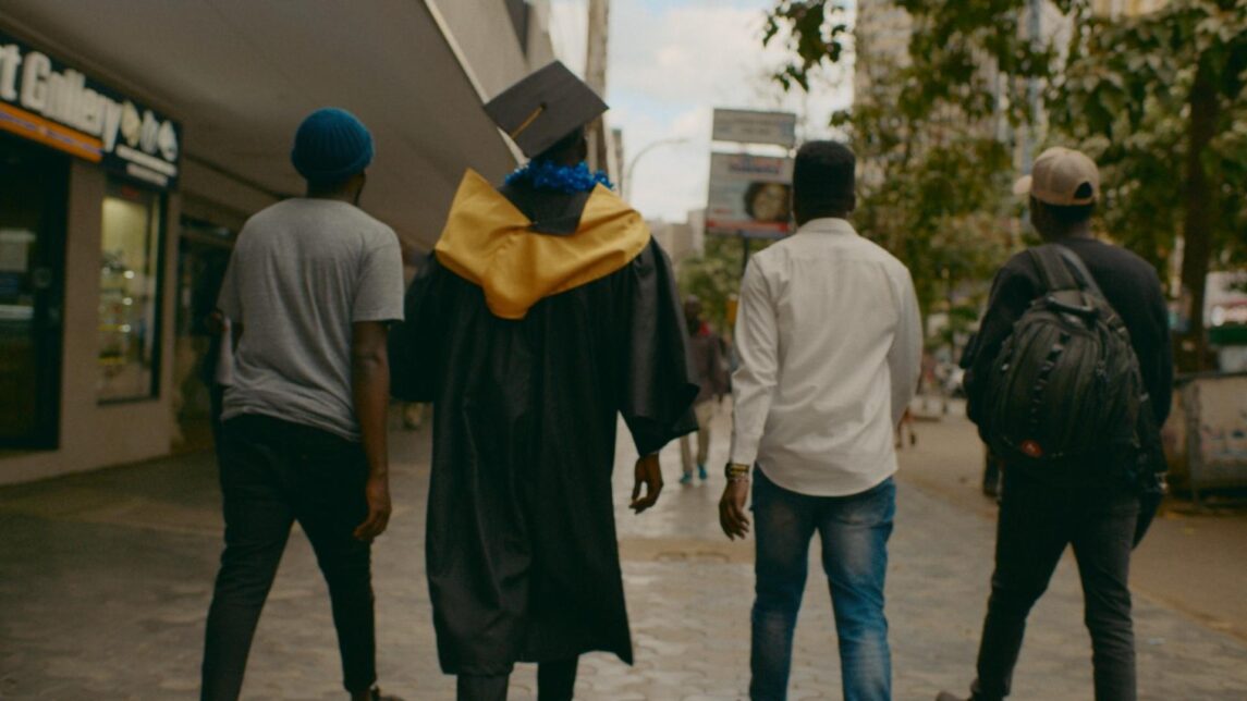 A film still from The Shadow Scholars shows the back of four Black men walking along a sidewalk. One of them wears a black graduation cap and gown along with a blue lei and gold stole.