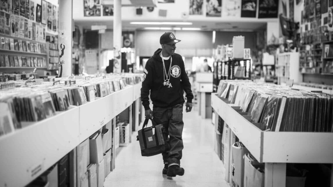 A black-and-white film still from The Sixth Borough shows a Black man walking down the aisle of a record store. He holds a bag in his hand and wears a Brooklyn Nets hat and hoodie along with a chain that reads: “RA”.