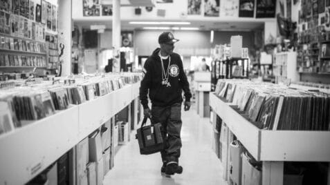 A black-and-white film still from The Sixth Borough shows a Black man walking down the aisle of a record store. He holds a bag in his hand and wears a Brooklyn Nets hat and hoodie along with a chain that reads: “RA”.