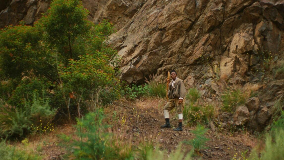 A film still from The Volcano Manifesto shows a Black man standing in the middle of the woods, leaning on a walking stick. His face is tense with concern, eyes shifted to the side and focused on something off screen. Behind him, large rocks rise out of frame into the sky, and he is surrounded by patches of trees,bushes, and other foliage.