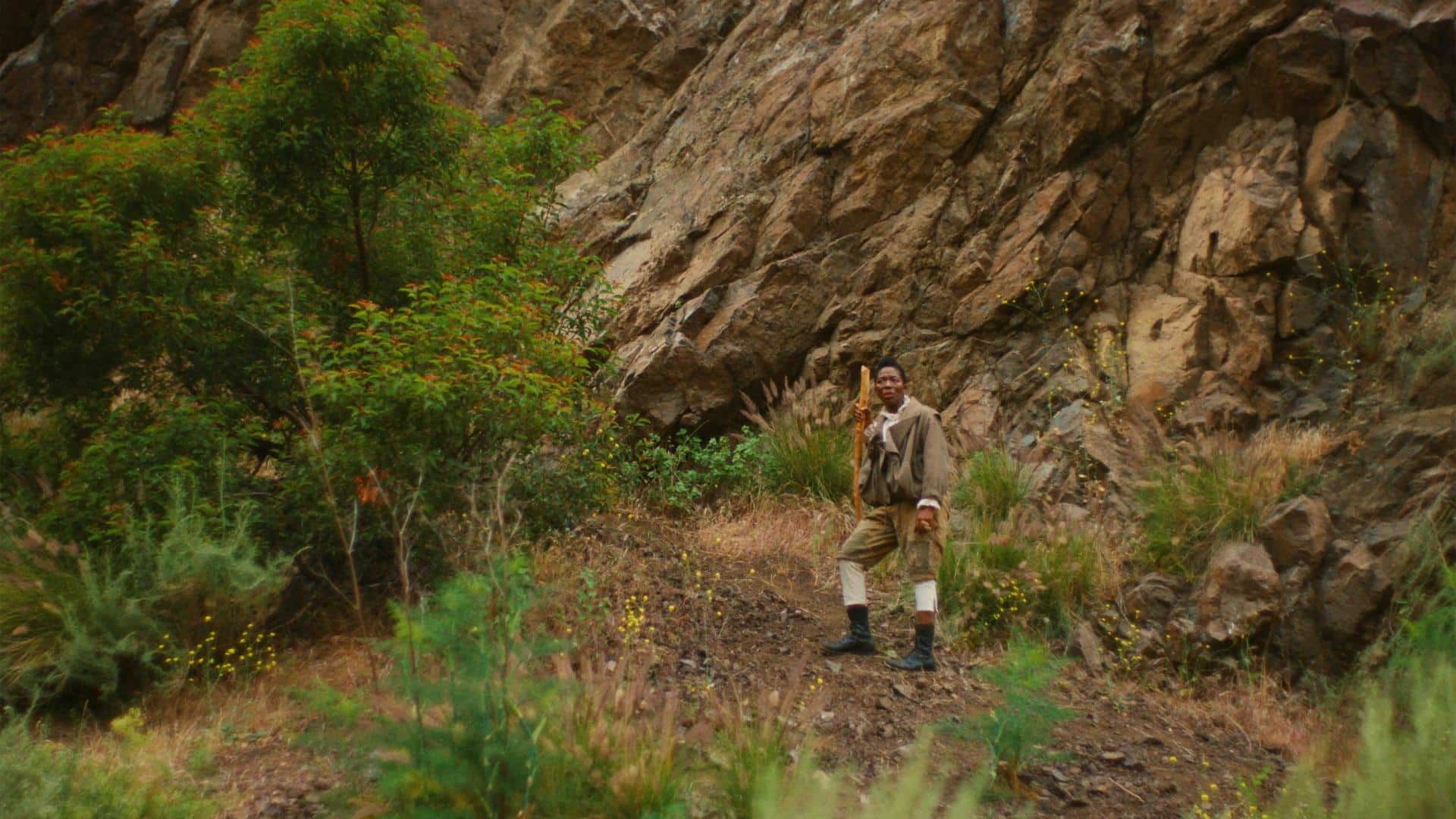 A film still from The Volcano Manifesto shows a Black man standing in the middle of the woods, leaning on a walking stick. His face is tense with concern, eyes shifted to the side and focused on something off screen. Behind him, large rocks rise out of frame into the sky, and he is surrounded by patches of trees,bushes, and other foliage.