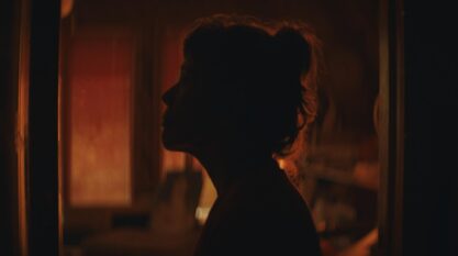 A film still from Tiger shows the silhouette of a woman’s side profile standing in a doorway. Her head is outlined by a red haze that comes from the room behind her, where the windows glow in a gradient that fades from red to orange.