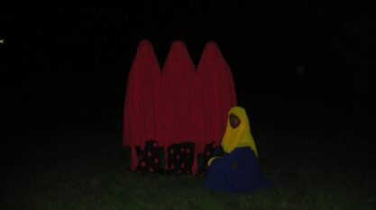 A film still from Twenty Three shows a dark field of grass. Emerging from the darkness stands three figures draped in red cloth over red and black pants. Sitting in front of them is a young woman wearing a long blue dress with a yellow hijab.