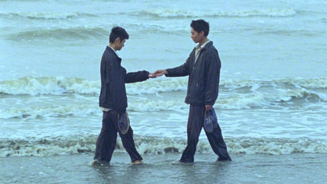 A film still from Viet and Nam shows two Asian men standing at the shoreline, facing each other. They wear matching navy blue jackets and pants, holding their shoes as the water washed over their feet. Their gaze is fixed on their hands, which together hold an unknown object.