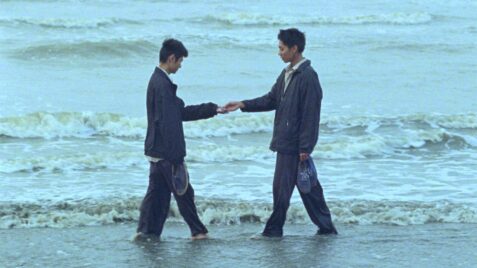 A film still from Viet and Nam shows two Asian men standing at the shoreline, facing each other. They wear matching navy blue jackets and pants, holding their shoes as the water washed over their feet. Their gaze is fixed on their hands, which together hold an unknown object.