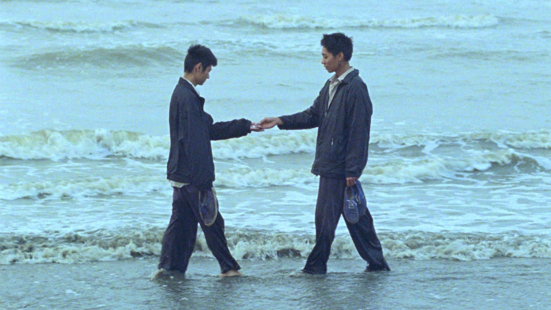 A film still from Viet and Nam shows two Asian men standing at the shoreline, facing each other. They wear matching navy blue jackets and pants, holding their shoes as the water washed over their feet. Their gaze is fixed on their hands, which together hold an unknown object.