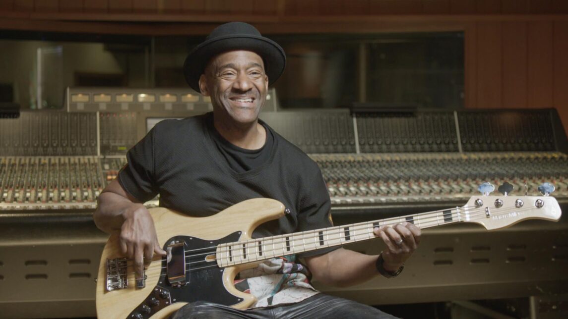 A film still from We Want the Funk shows a Black man seated in a music recording studio, holding a guitar. He smiles excitedly.