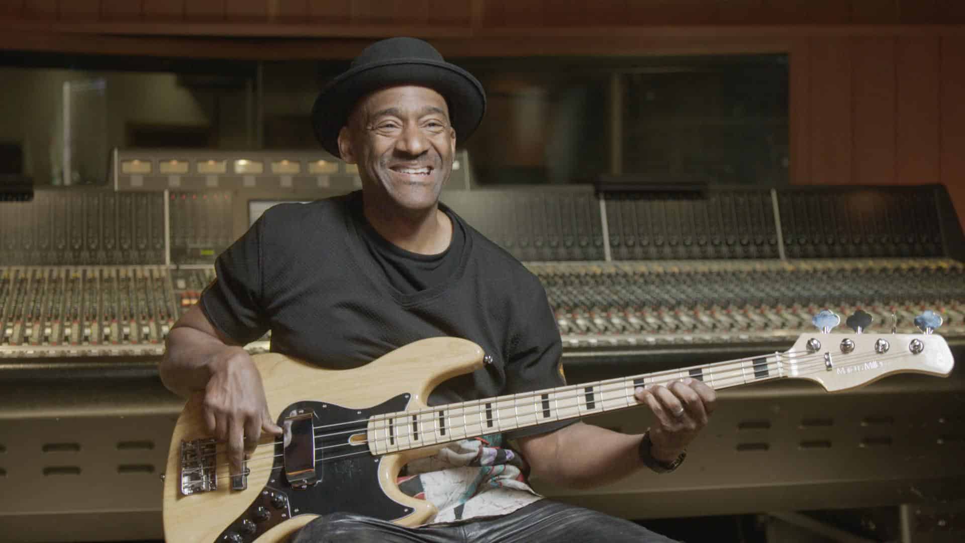 A film still from We Want the Funk shows a Black man seated in a music recording studio, holding a guitar. He smiles excitedly.