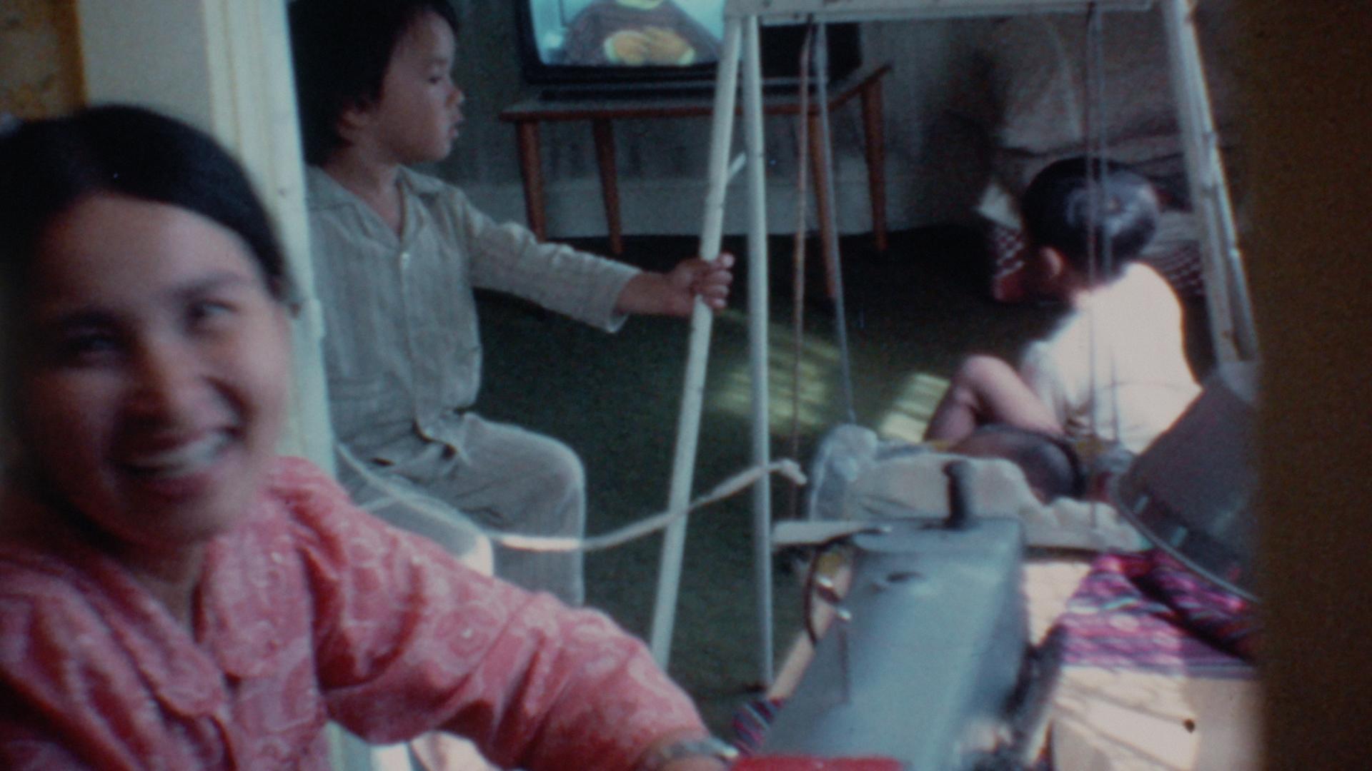 A film still from We Were the Scenery shows an Asian woman smiling as she is captured in motion in a door frame. Behind her, two Asian children rest. One child looks at the other, who is lying on a small swing and focused on a TV screen.
