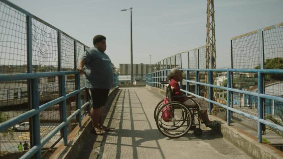 A film still from White House shows an older Black woman in a wheelchair looking out from an overpass above a road. Behind her, a young Black man leans against the opposite fence of the overpass.