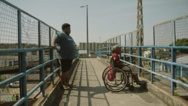 A film still from White House shows an older Black woman in a wheelchair looking out from an overpass above a road. Behind her, a young Black man leans against the opposite fence of the overpass.