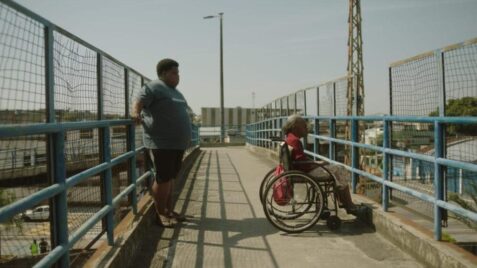 A film still from White House shows an older Black woman in a wheelchair looking out from an overpass above a road. Behind her, a young Black man leans against the opposite fence of the overpass.