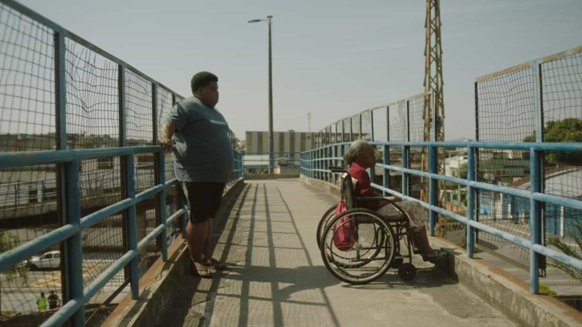 A film still from White House shows an older Black woman in a wheelchair looking out from an overpass above a road. Behind her, a young Black man leans against the opposite fence of the overpass.
