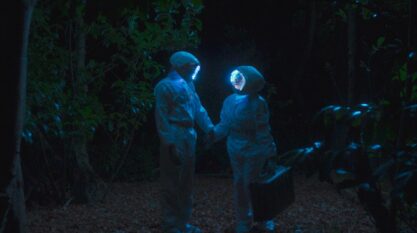 A film still from Why the Sun Moon Live in the Sky shows two astronauts standing in the middle of the woods, holding hands and sharing a gaze. Their helmets emit a soft blue glow that lights the surrounding darkness.