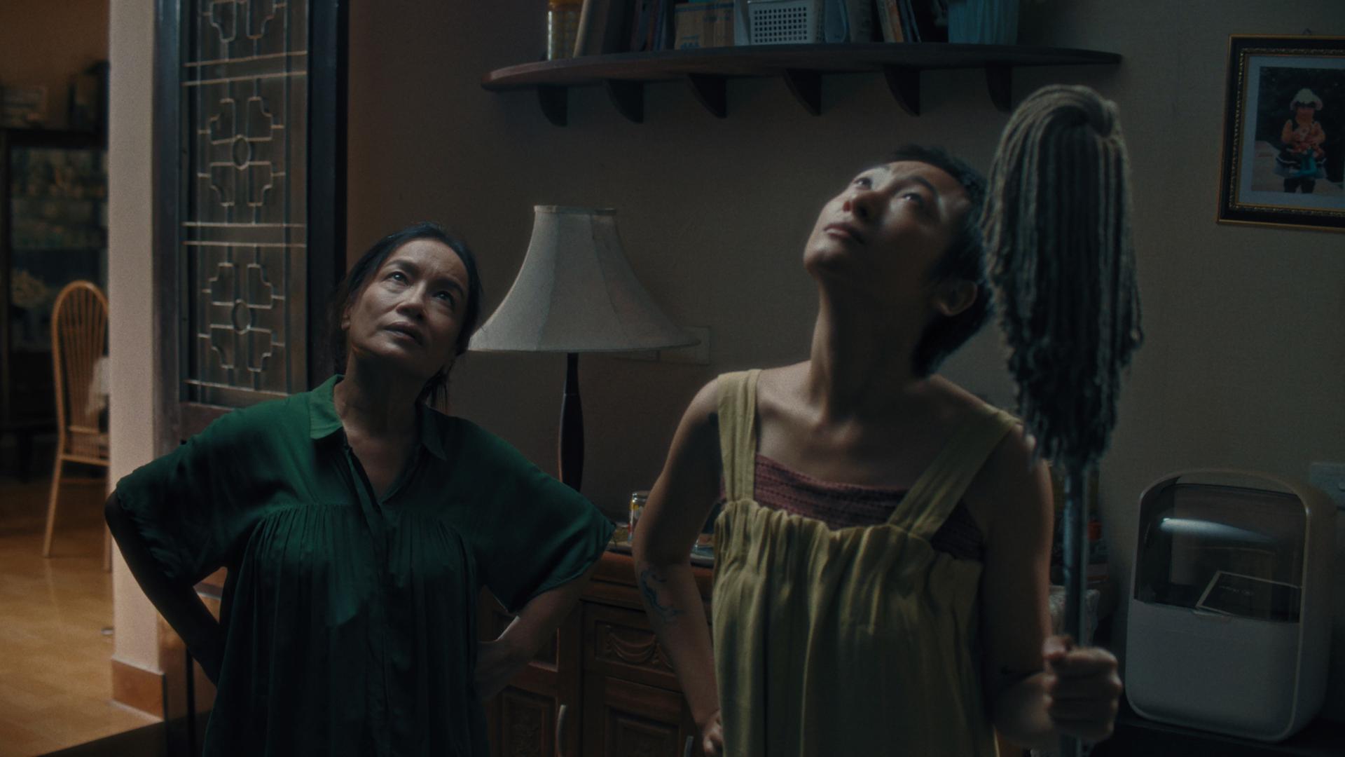 A film still from Don’t Cry Butterfly shows two Asian people standing side by side looking up at presumably the ceiling of a home. The person on the right is folding a mop.