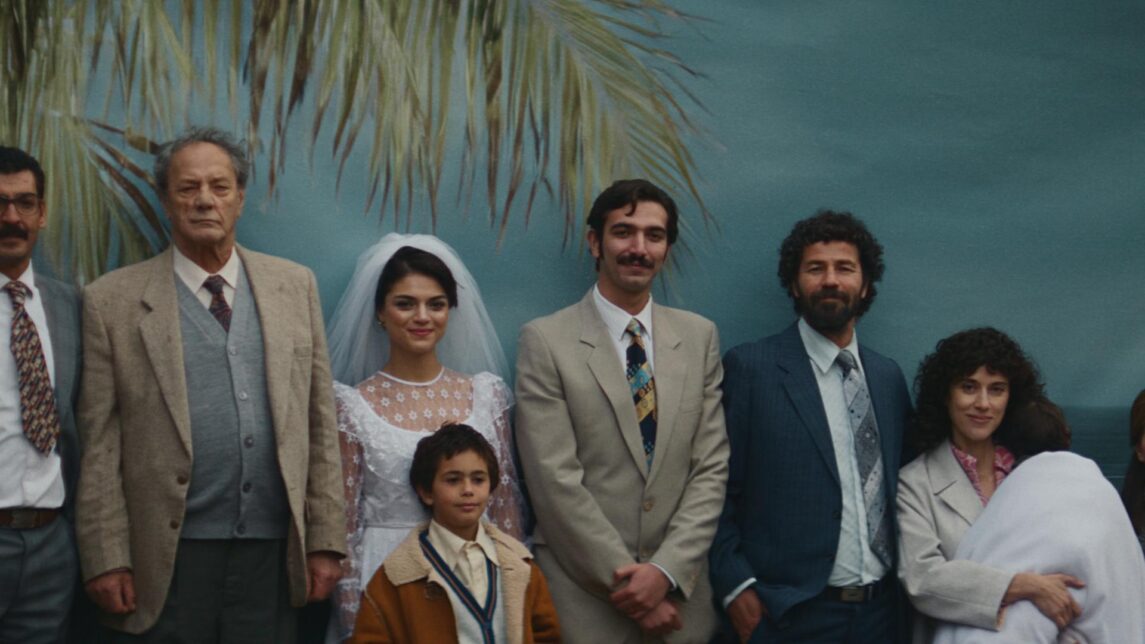 A film still from All That’s Left of You shows a group of Palestinians posing in front of a blue backdrop with a painted palm tree. At the center stands a woman in a white wedding dress and veil, with a young boy in front of her. Two men in suits stand to her left, and two more men in suits stand to her right. A woman holding presumably a baby wrapped in a white cloth stands nearby, along with another woman in a fur coat.