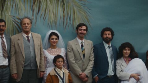 A film still from All That’s Left of You shows a group of Palestinians posing in front of a blue backdrop with a painted palm tree. At the center stands a woman in a white wedding dress and veil, with a young boy in front of her. Two men in suits stand to her left, and two more men in suits stand to her right. A woman holding presumably a baby wrapped in a white cloth stands nearby, along with another woman in a fur coat.