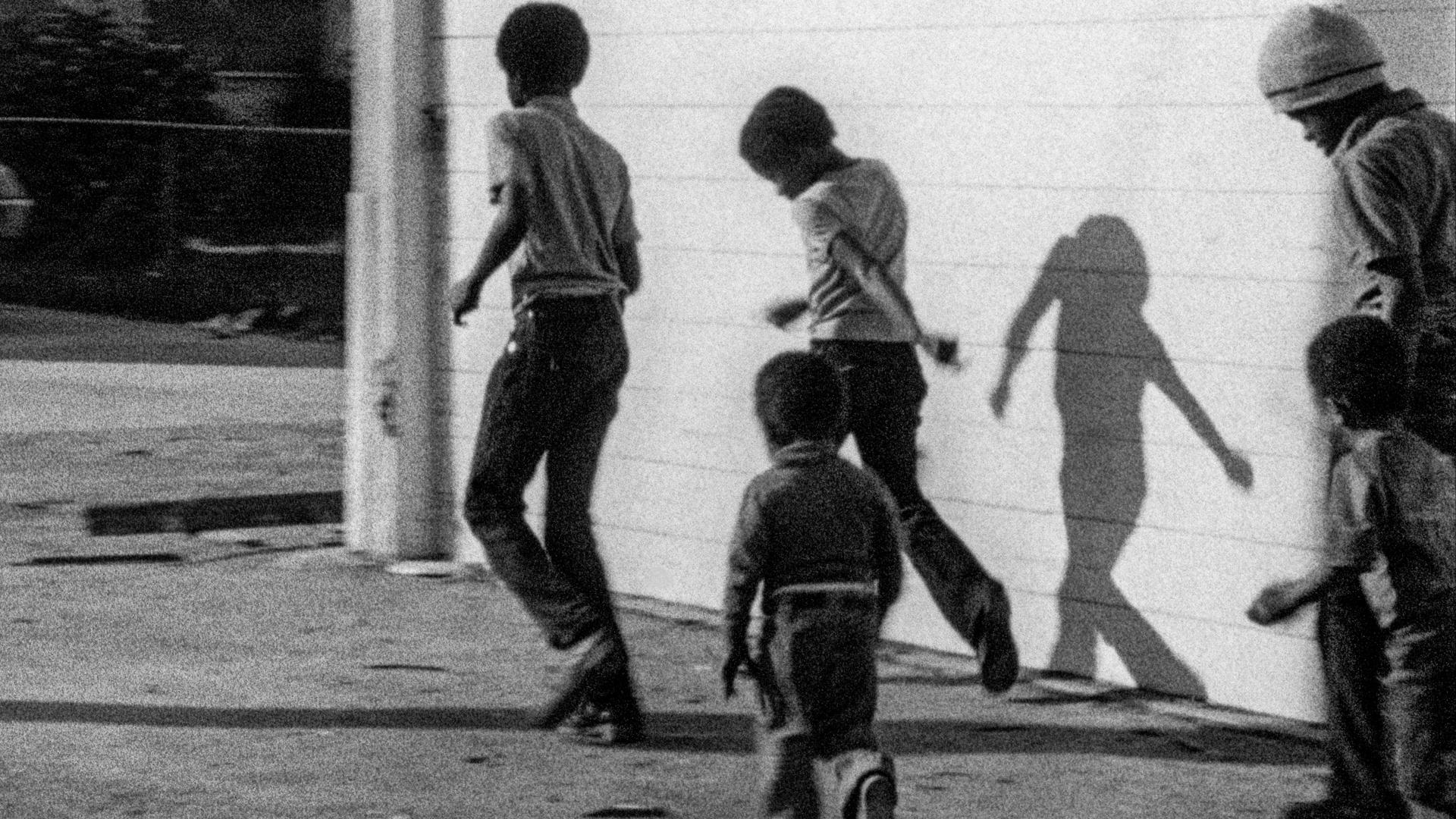 A black-and-white film still from Killer of Sheep shows a group of young Black boys walking past the side of a house.