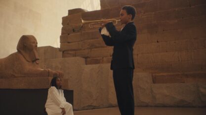 A film still from Untitled (How High the Moon) shows a young Black boy in a suit playing the trumpet. In front of him, a young Black girl dressed in all white is looking up at him. Behind her is a sphinx positioned in front of presumably the side of a pyramid.