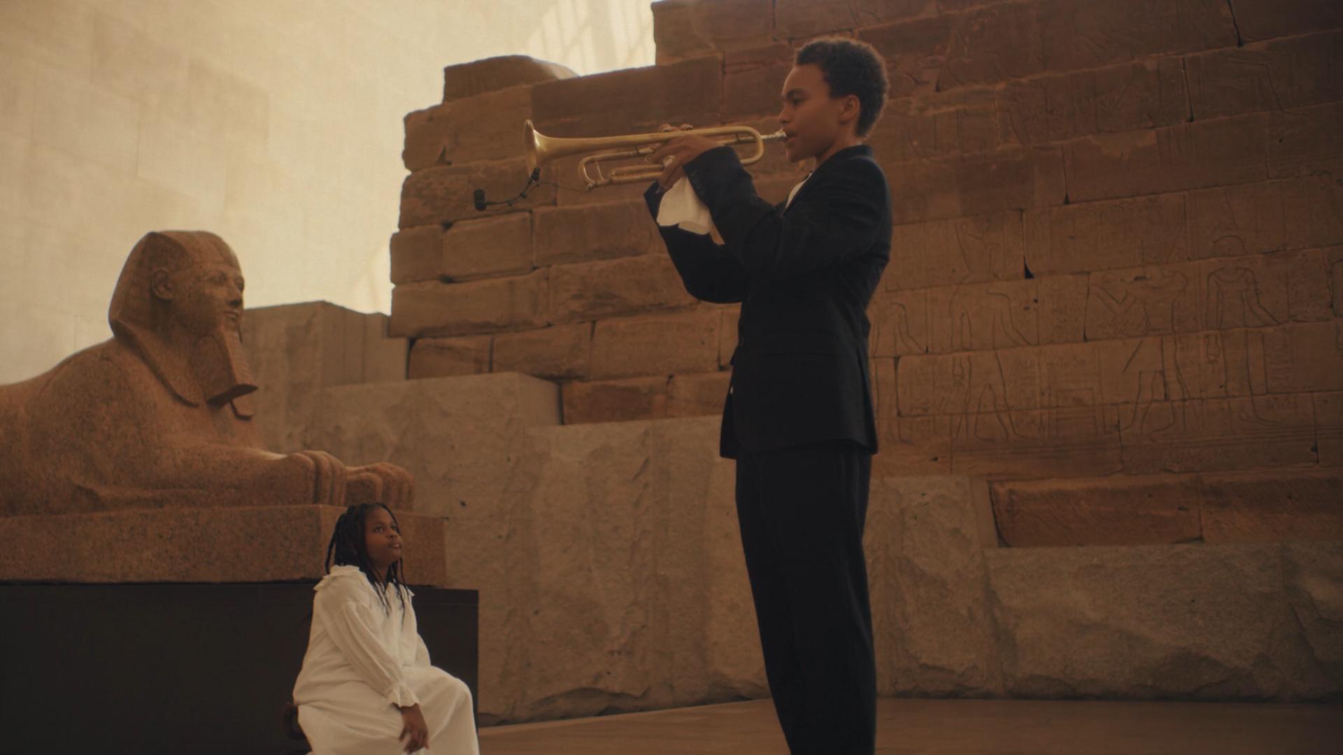 A film still from Untitled (How High the Moon) shows a young Black boy in a suit playing the trumpet. In front of him, a young Black girl dressed in all white is looking up at him. Behind her is a sphinx positioned in front of presumably the side of a pyramid.