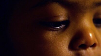 A film still from Axel shows the face of a Black child emerging from darkness. The child’s eyes are fixed down.