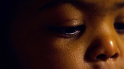 A film still from Axel shows the face of a Black child emerging from darkness. The child’s eyes are fixed down.