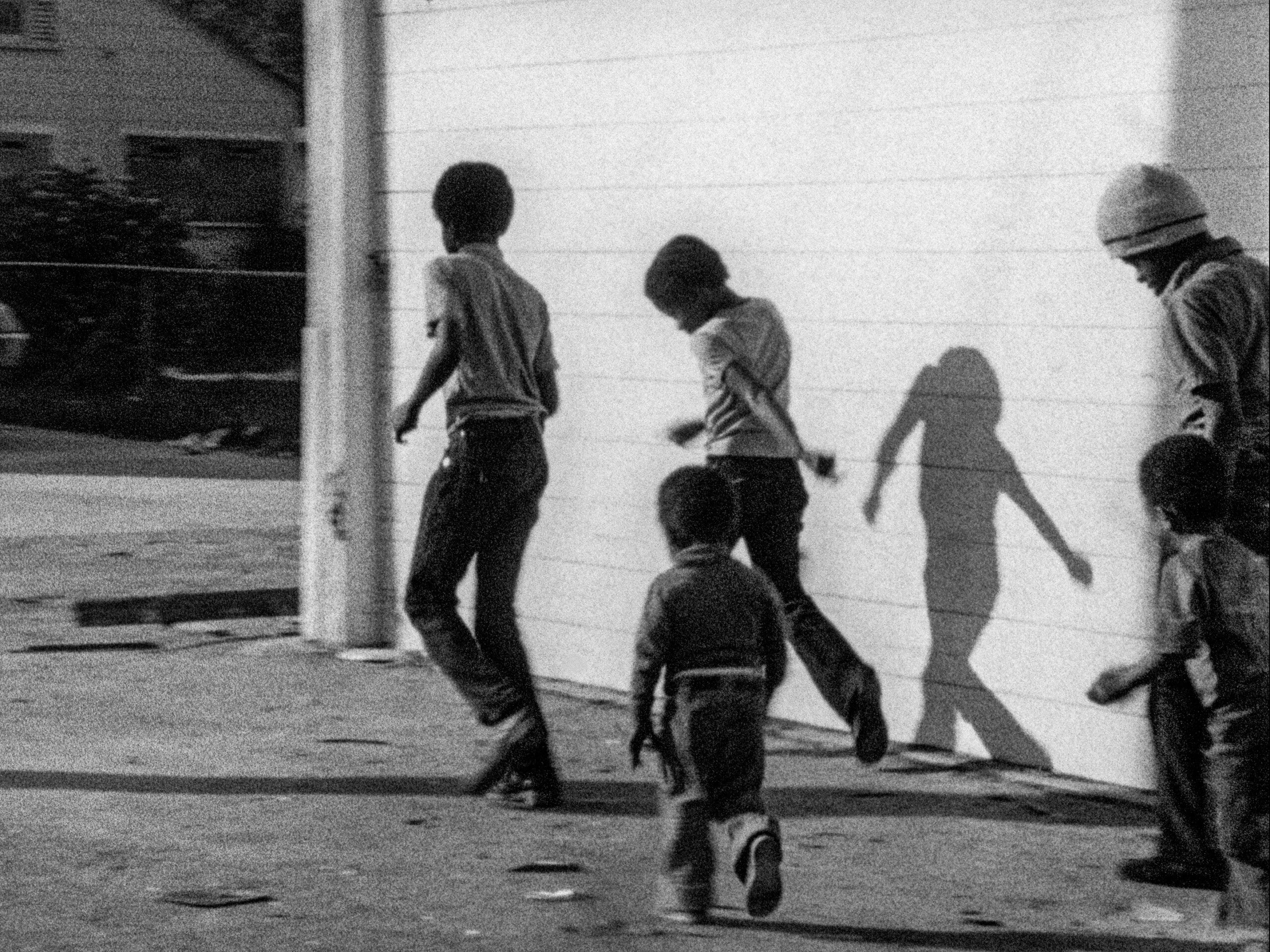 A black-and-white film still from Killer of Sheep shows a group of young Black boys walking past the side of a house.