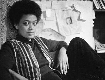 A black-and-white film still from TCB – The Toni Cade Bambara School of Organizing shows Toni Cade Bambara leaning against a paisley-printed curtain, gazing directly at the camera. She wears an afro and an open striped vest over a long-sleeve top. Behind her is a board with papers pinned to it and a shelf with books.