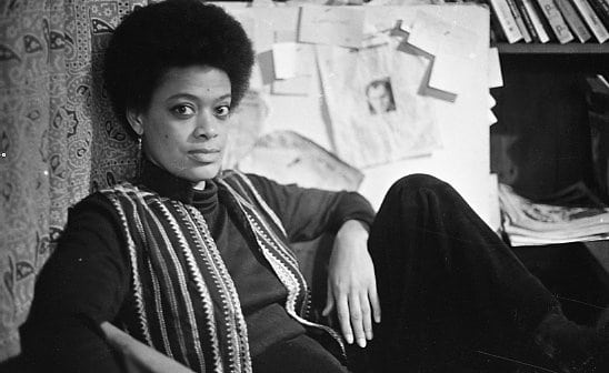 A black-and-white film still from TCB – The Toni Cade Bambara School of Organizing shows Toni Cade Bambara leaning against a paisley-printed curtain, gazing directly at the camera. She wears an afro and an open striped vest over a long-sleeve top. Behind her is a board with papers pinned to it and a shelf with books.