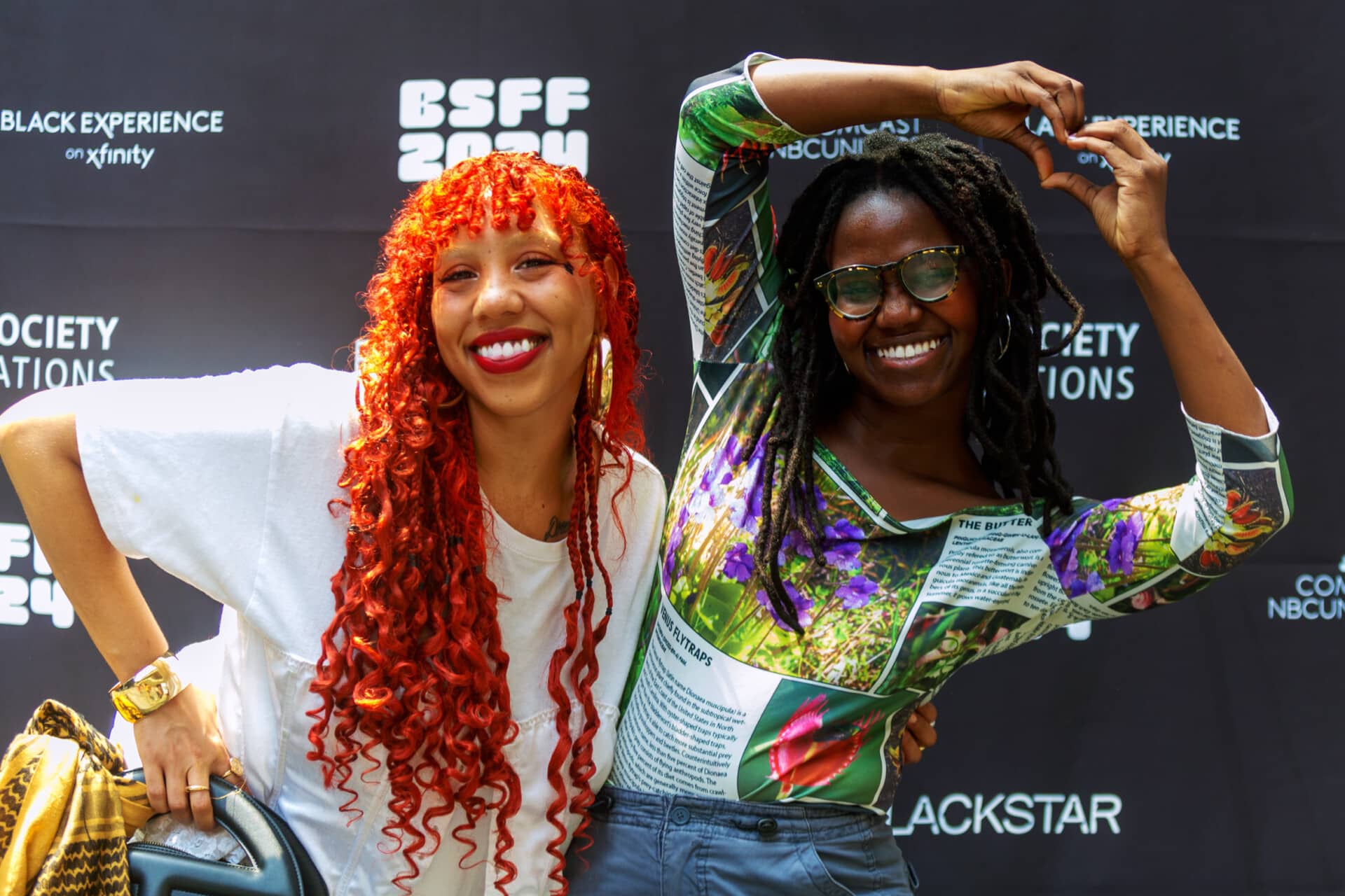 A photo of two people posing in front of the step-and-repeat at the 2024 BlackStar Film Festival. They are smiling. One has their hands raised into a heart shape.