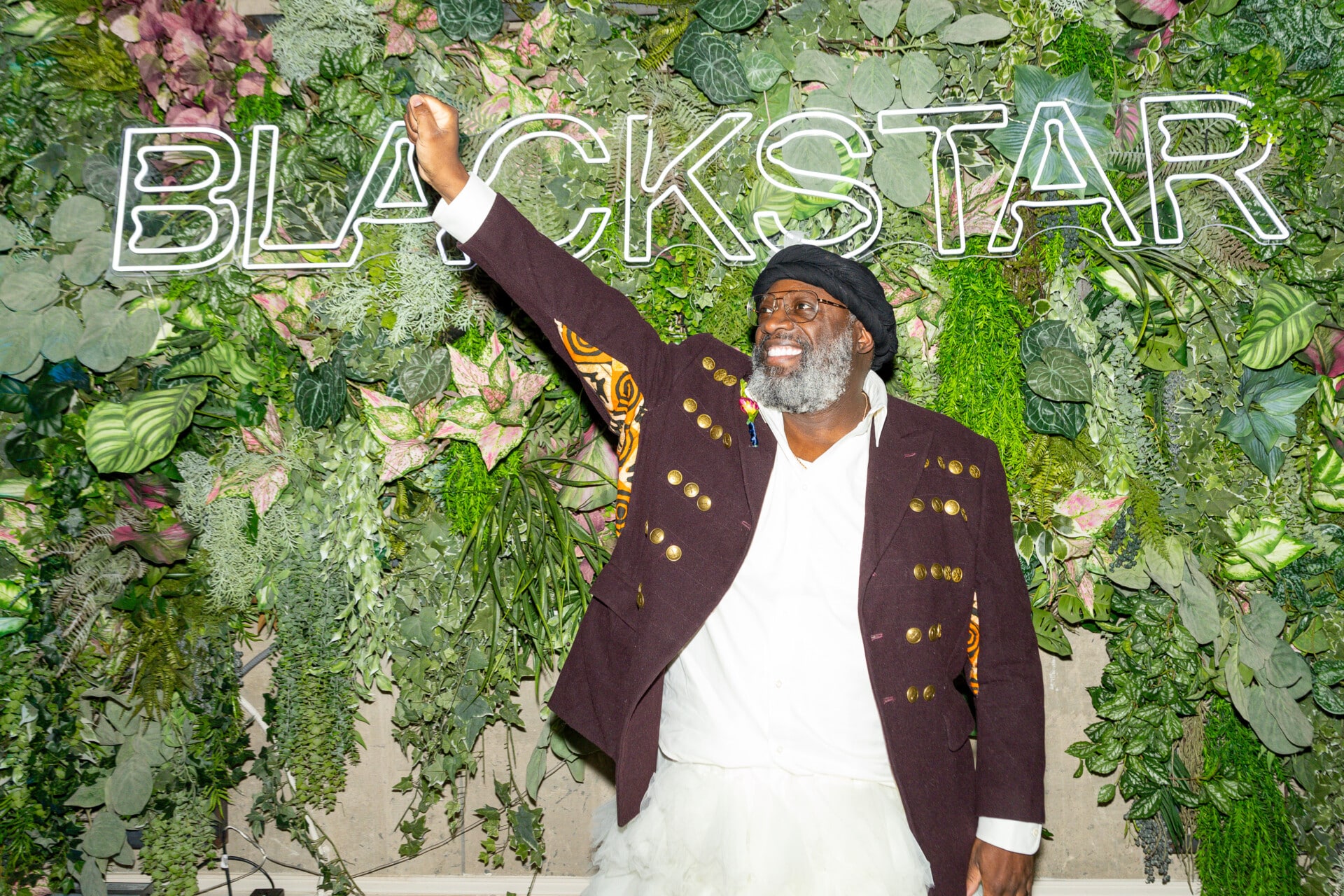 A photo from the 2024 BlackStar Luminary Gala of a Black man standing in front of a BlackStar sign, his fist is raised in the air and he is smiling.