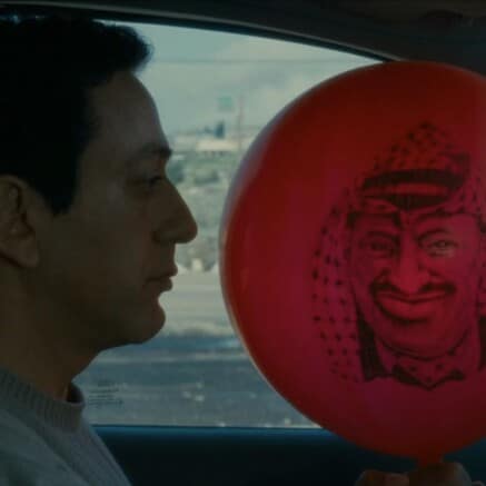 A film still from Divine Intervention shows a close up of a man in car, he has a somber expression and is holding a big red ballon with a person's face printed on it in black. The person on the ballon has a scarf on their head and is smiling menacingly.