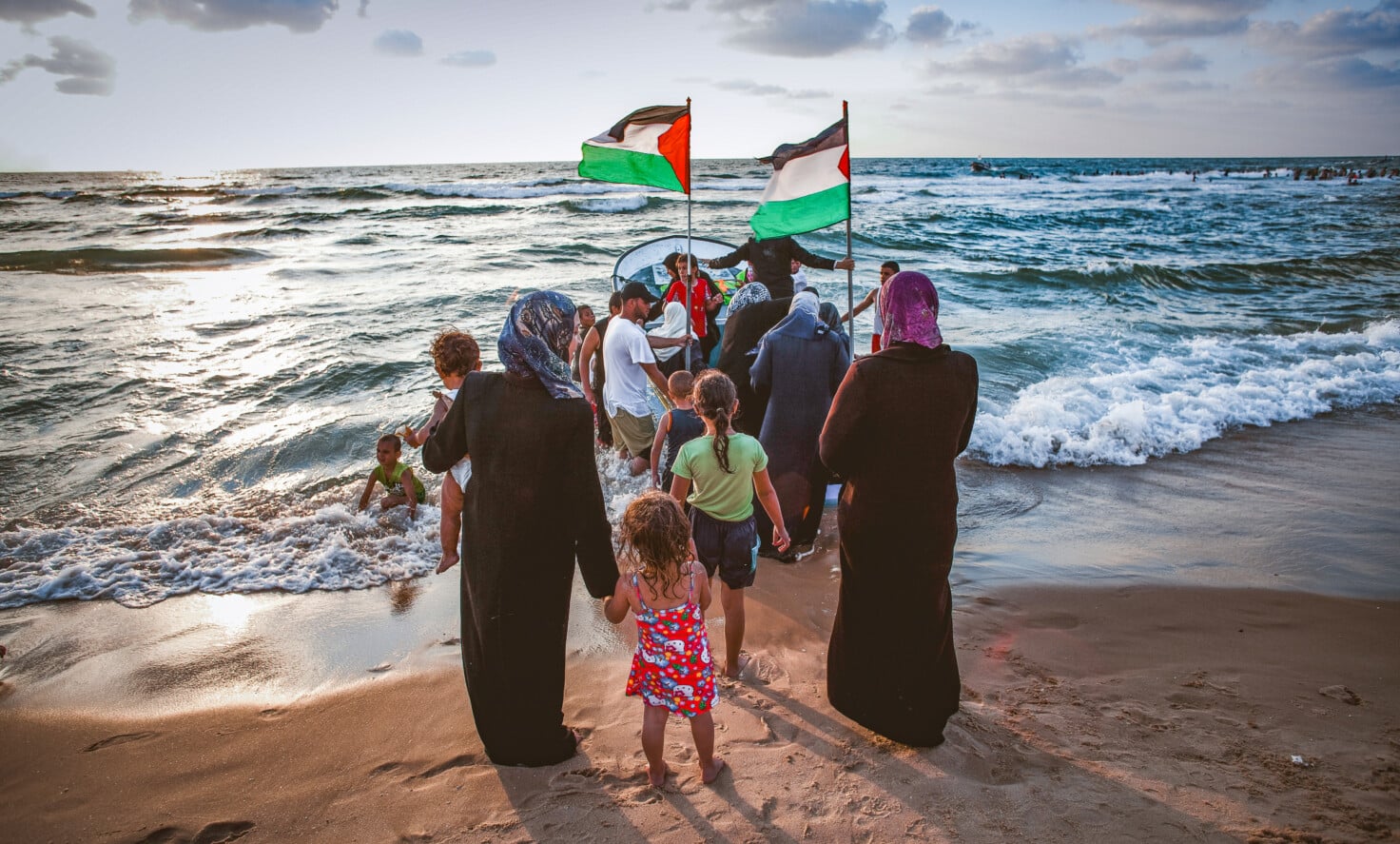 Arab women wearing headscarves and children are standing on a beach waiting to get on a boat. The boat has two Palestinian flags hanging high on poles.