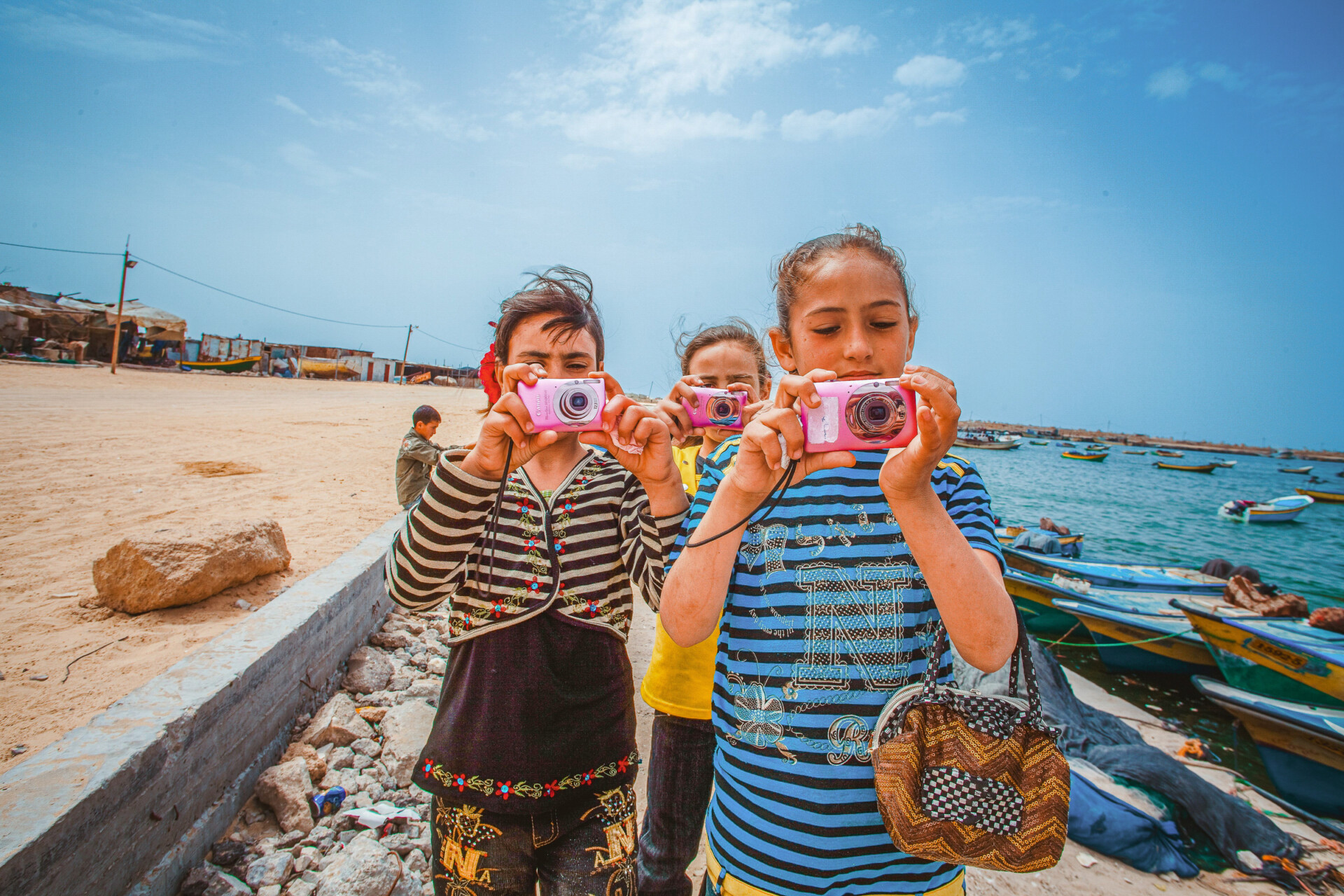 Three Palestinian girls hold pink point-and-shoot cameras directly at the person taking their photograph. Two of them are wearing colorful striped shirts.
