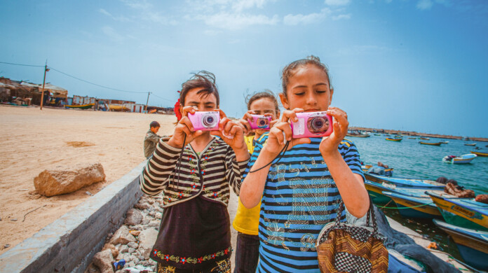 Three Palestinian girls hold pink point-and-shoot cameras directly at the person taking their photograph. Two of them are wearing colorful striped shirts.