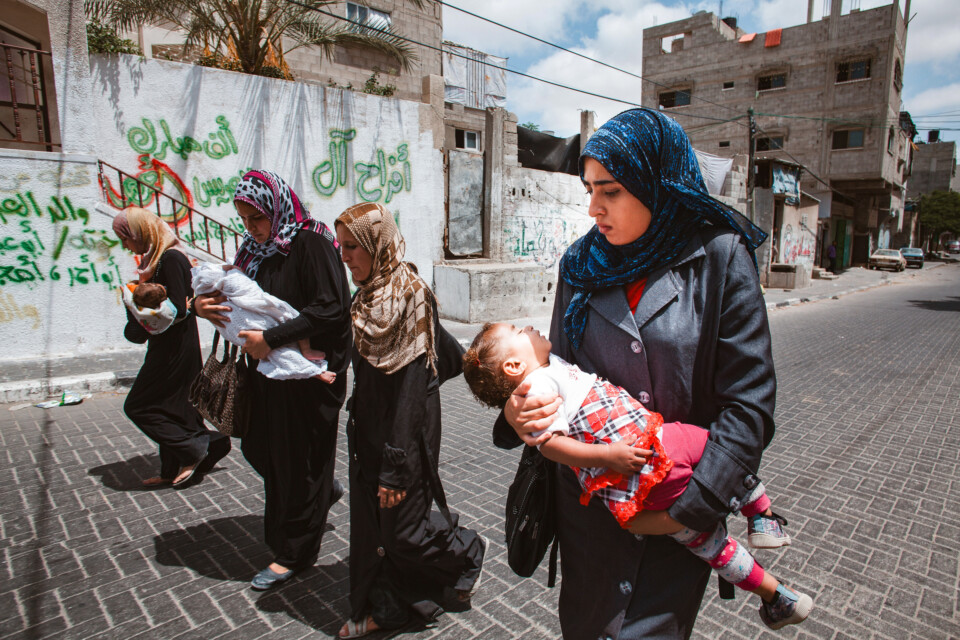 Four hijabi women walk alongside each other with their heads hung. Three of them hold babies in their arms.