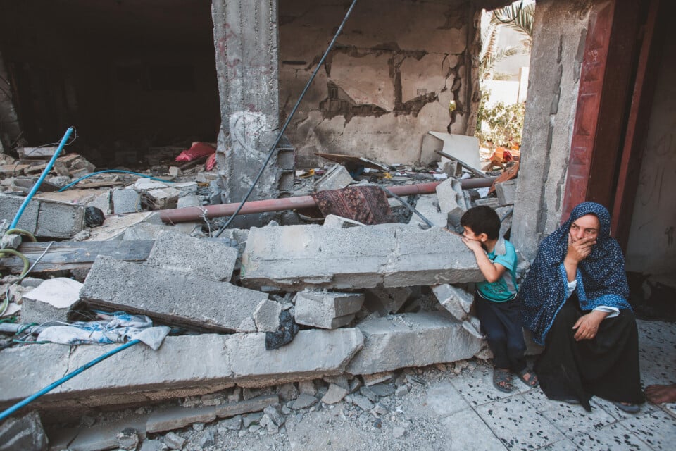 A woman in a blue polka dot hijab and a young boy wearing a teal shirt and sandals sit in the concrete ruins of their decimated home.