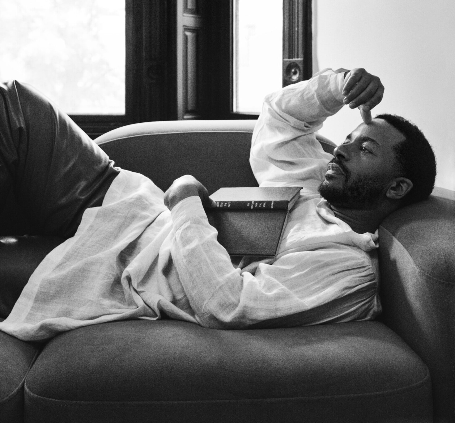 André Holland reclines on a couch, looking pensively into the distance. His arm is above his head, and a sliver of a book can be seen resting on his chest. The photo is in black and white.