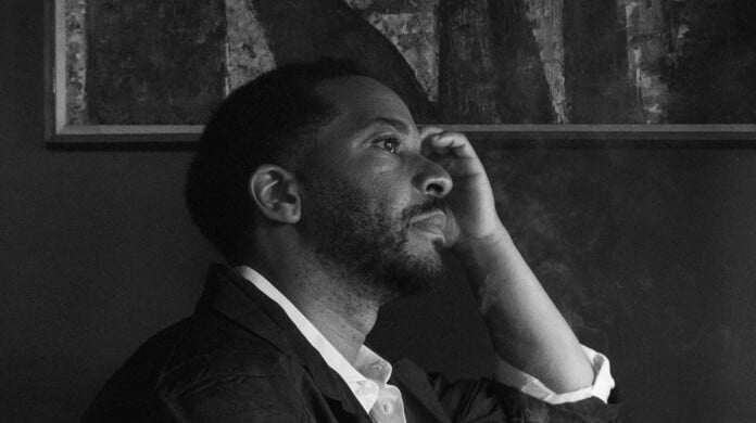 A picture of André Holland, in profile, looking to the right with his left hand on his head and his right hand holding a small tray with smoke rising from it.