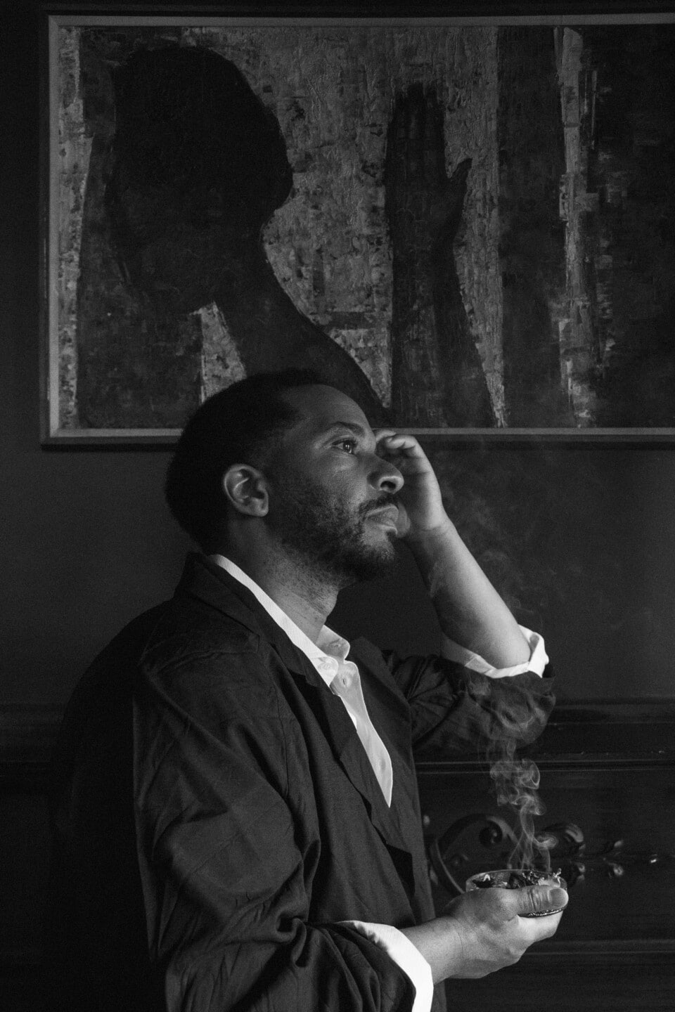 A picture of André Holland, in profile, looking to the right with his left hand on his head and his right hand holding a small tray with smoke rising from it.